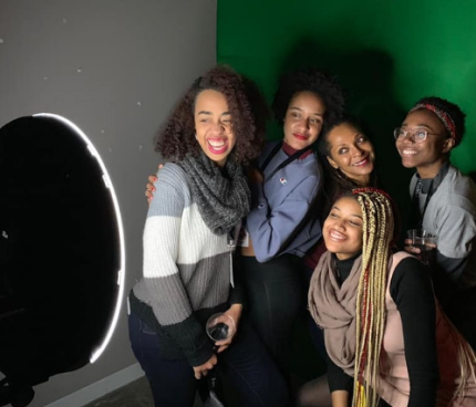 Five Black women pose in front of a green screen, a large ring light pointing at them.
