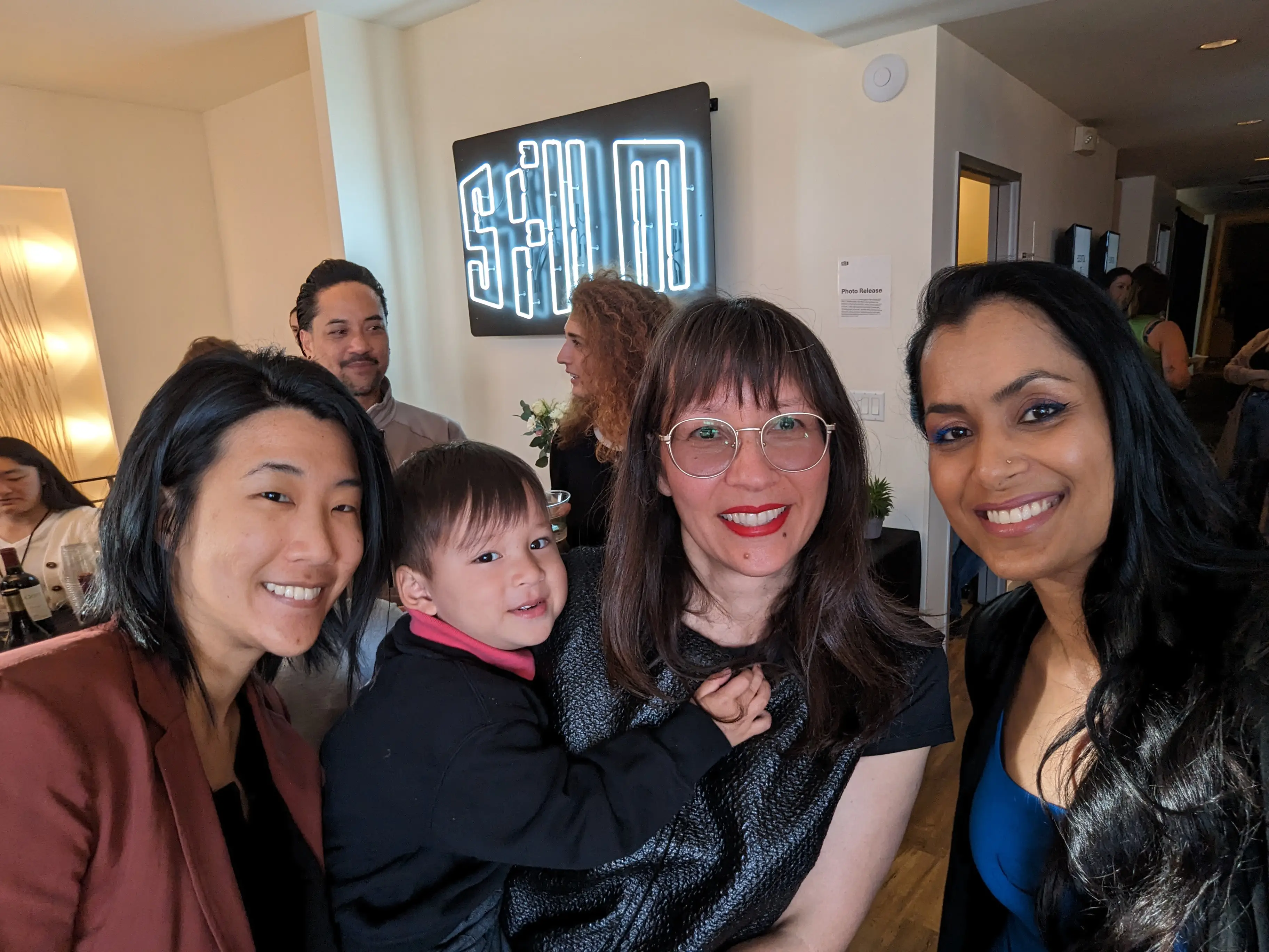 Three BGDM members and a toddler smile in front of the SFFILM neon sign. From left to right, Asian Betsy Tsai, Asian Ursula Liang and her son, and another woman of color.