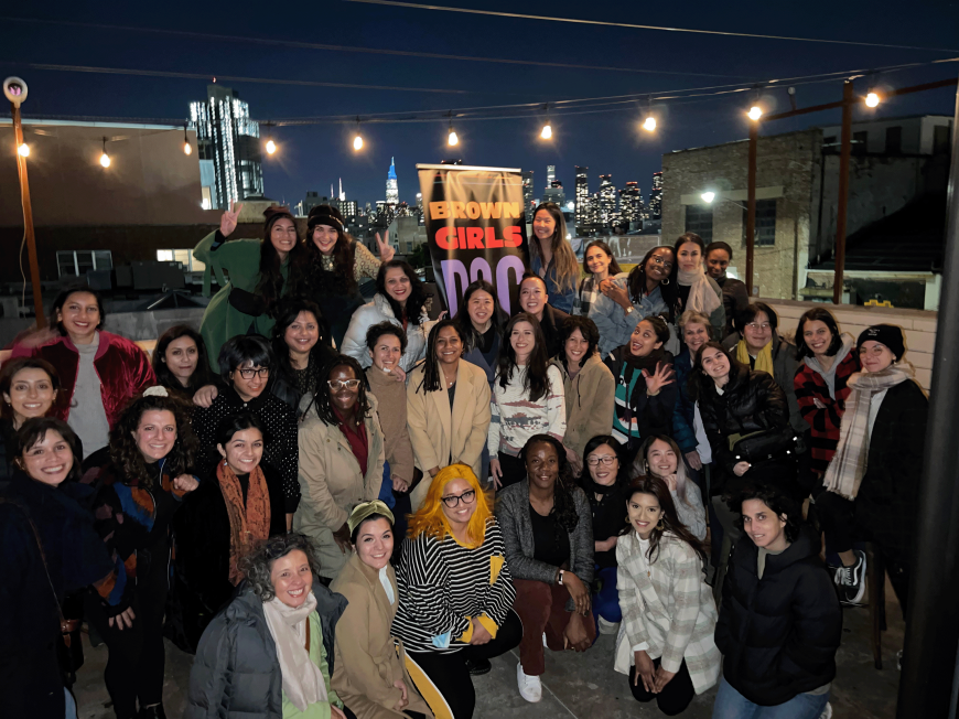 A large group of women and nonbinary people of color pose, with a sign that says 'Brown Girls Doc Mafia', the Manhattan skyline behind them.