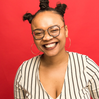 Daquisha is a Black woman with rounded glasses, dark lipstick,a striped blouse and her hair in three bantu knots.