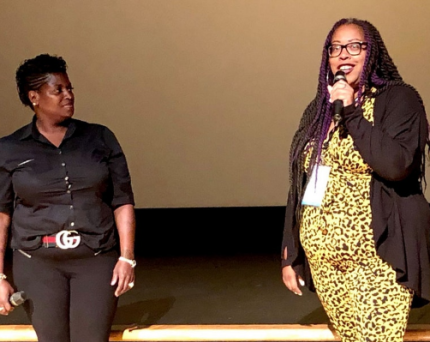 Two Black women with microphones speaking on a stage, one with short braided back hair and a black button up, the other with long purple twists and a yellow cheetah print dress.