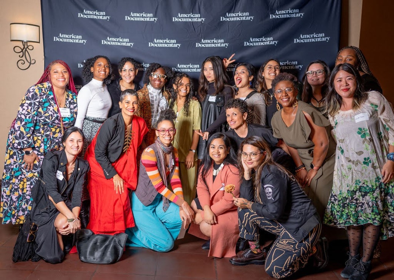 A large group of BIPOC women and nonbinary people gather for a photo in front  of a navy blue backdrop with the words 