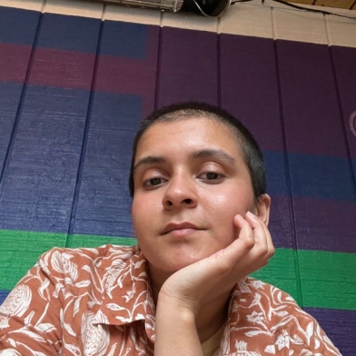 Rajvi is an trans and nonbinary Indian person with a buzzcut and patterned orange and white shirt.