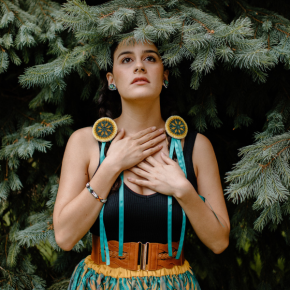 Victoria is an Indigenous Bolivian-American women with long dark brown braids, beaded hair pieces an a teal skirt.