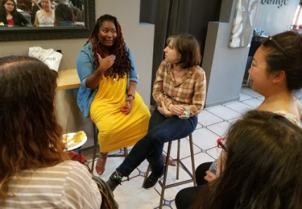 A Black woman with curly dreadlocks in yellow speaks to four other women of color.