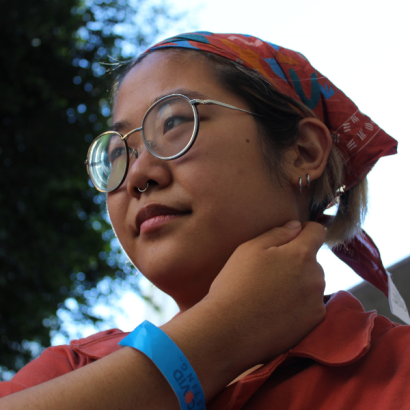 Jireh is an Asian-American person with round glasses, double-pierced ears and a red bandana pulling back their hair.