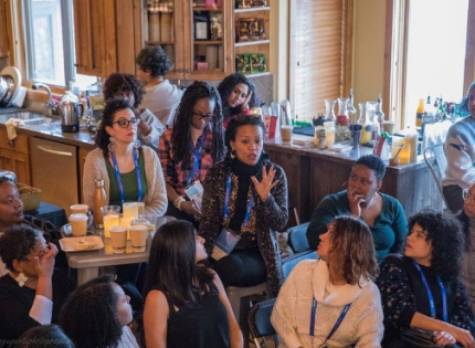 Sitting at a table in a large dining room, a Black woman speaks to a crowd of people.