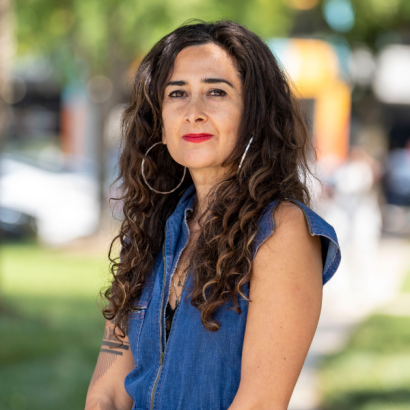 Sahar is an Iranian American woman with long, voluminous curly hair, large hoop earrings and a blue sleeveless denim jumpsuit.