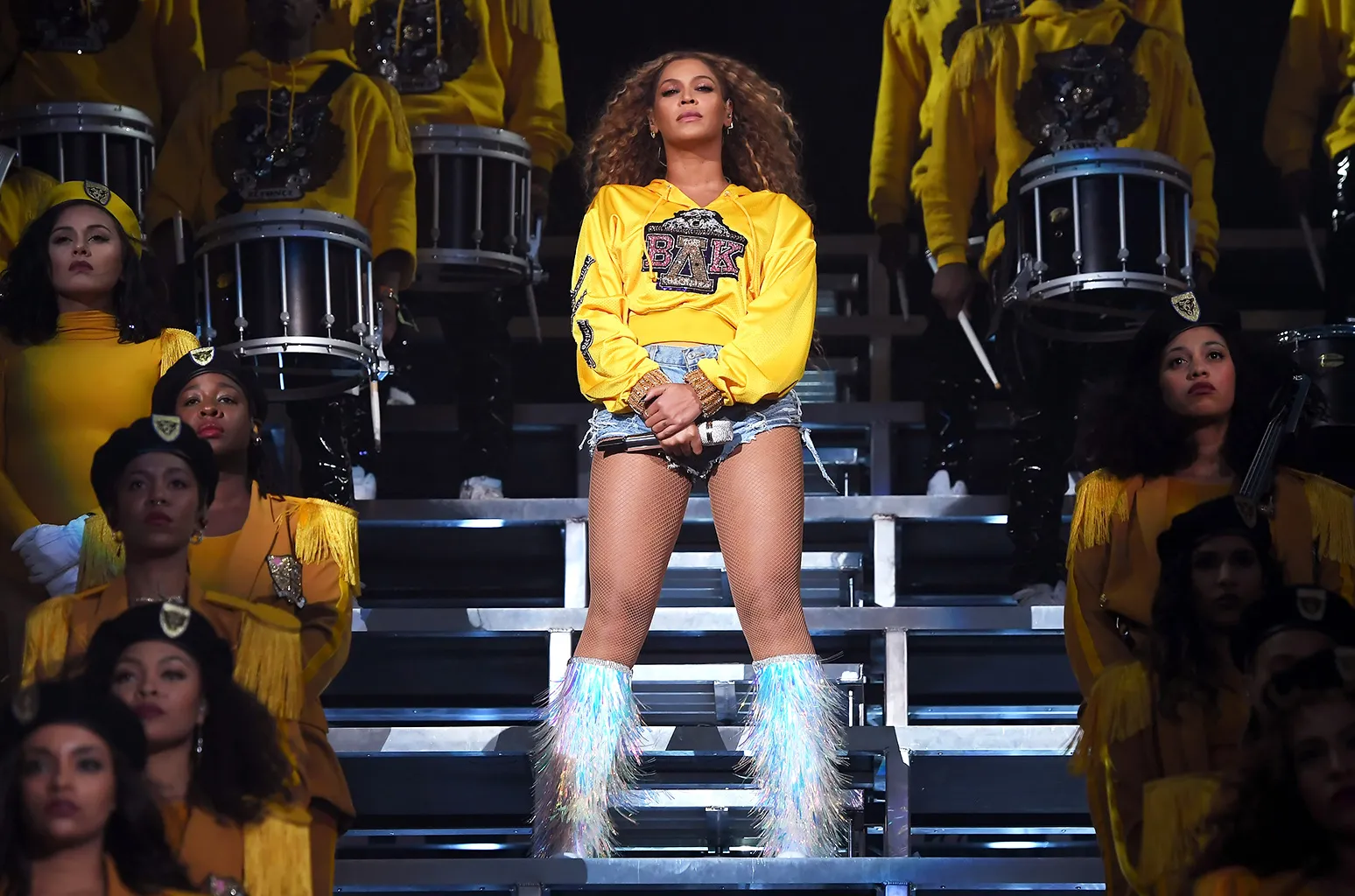 Beyonce stands on bleachers, surrounded by dancers, in a yellow sweatshirt, small jean shorts and holographic fringe boots.