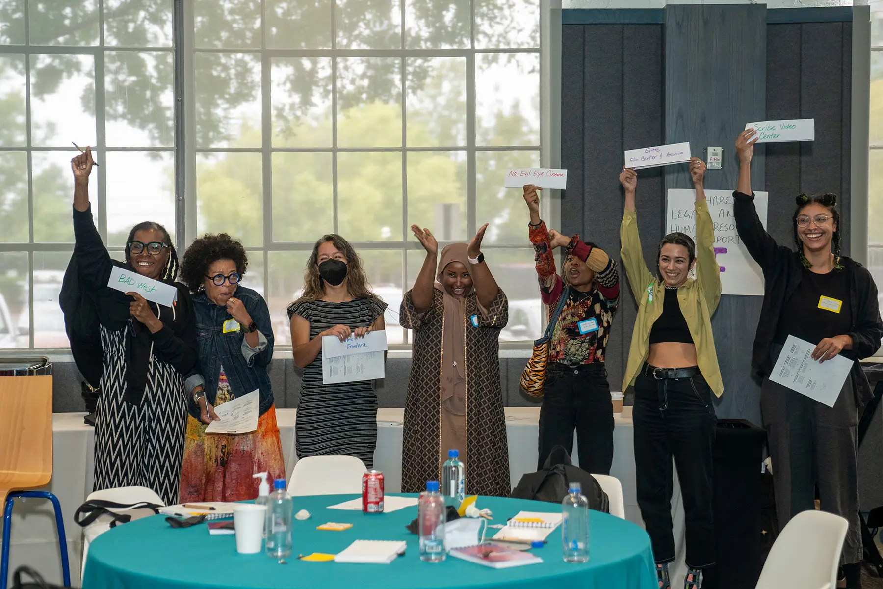 A group of women of color hold up pieces of paper, cheering, as they stand around the table at the Color Congress Convening
