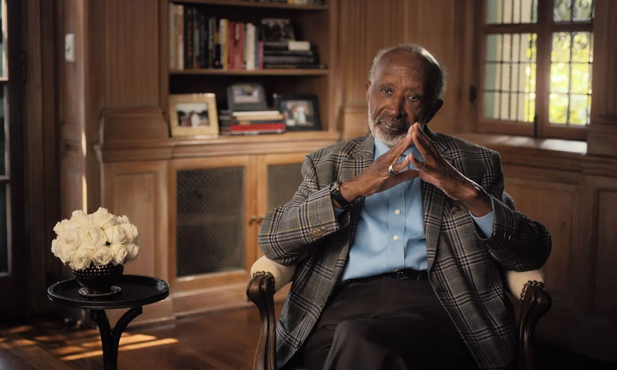 Clarence Avant, a bald Black man with a white beard and a suit, sits in a chair in front of a cozy wood-paneled living room.