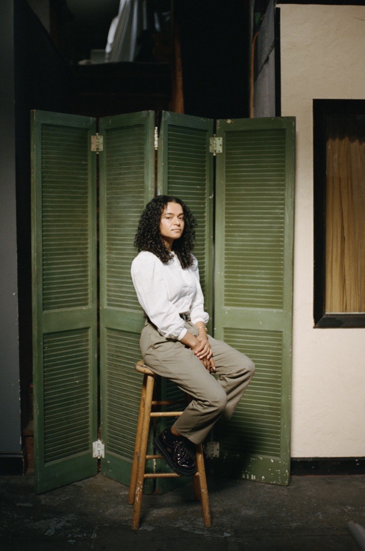 Aurora is a Black and Bangladeshi woman with curly hair and a white blouse. She sits on a stool in front of a green folding door.