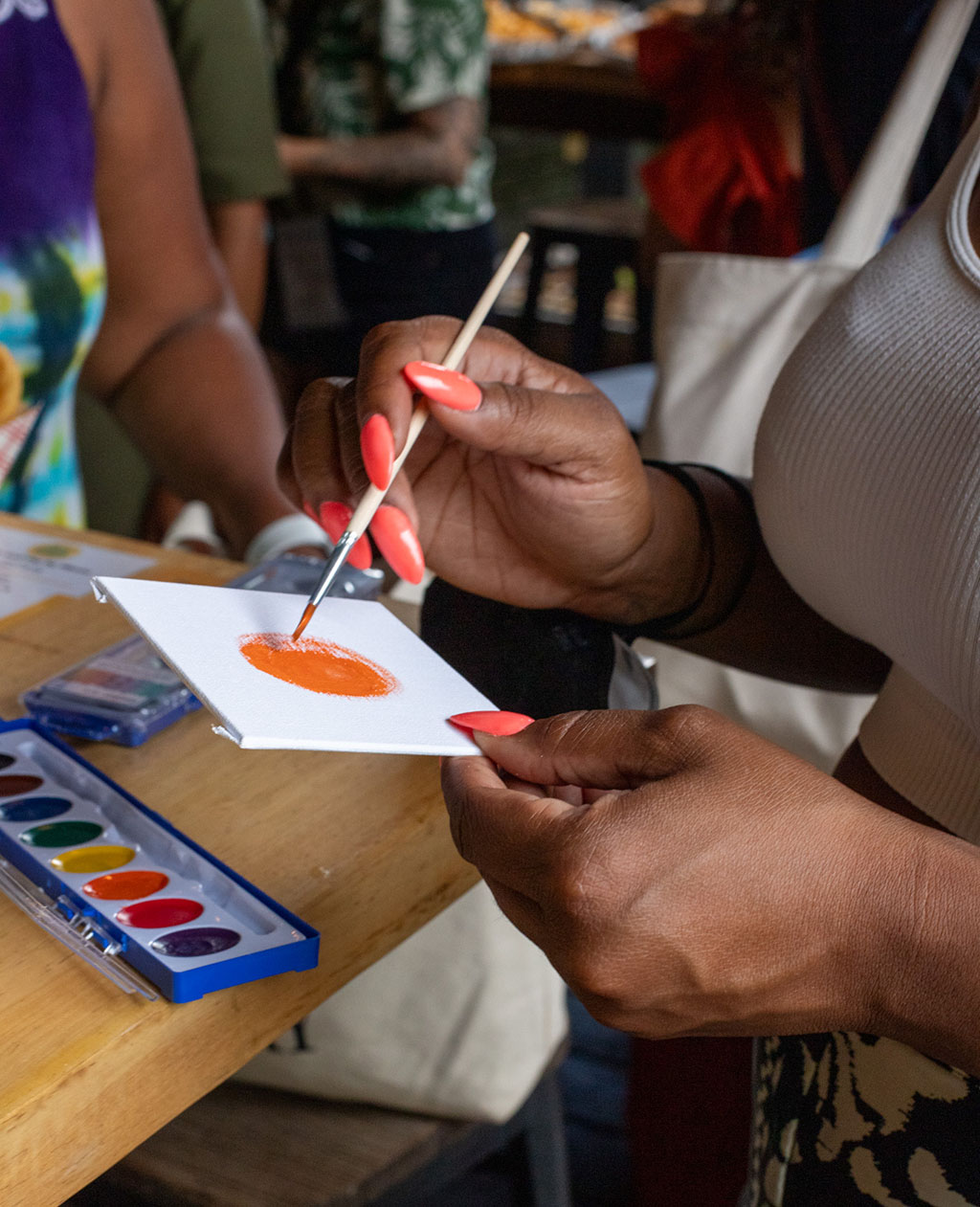 A woman of color paints an orange circle on a square white canvas with watercolor.