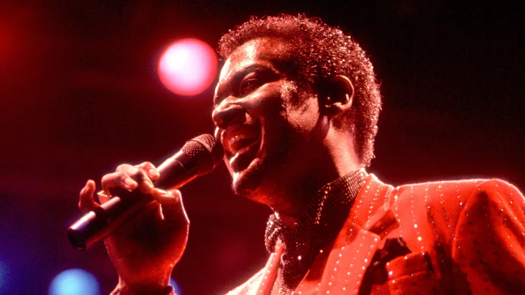 Under red stage lights, Luther Vandross, a Black man with short hair and a suit, sings passionately into a microphone.