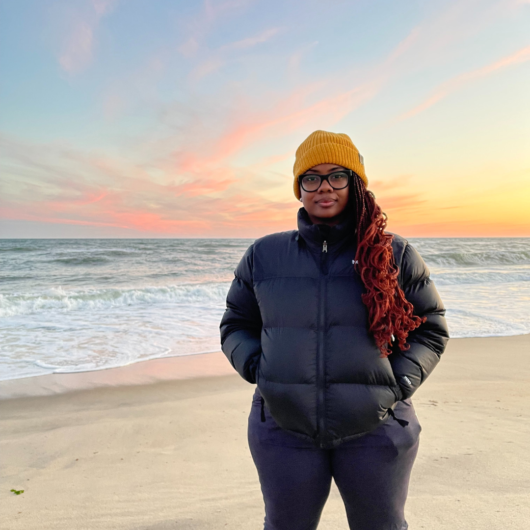 Brittany Ferrell is a Black woman with long red braids, a yellow beanie and a large black puffer jacket. She stands on a beach at sunset.