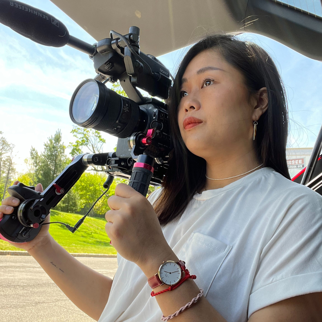 Chinese Xinyan, a women with medium-length black hair and a white shirt, holds a large cine camera.