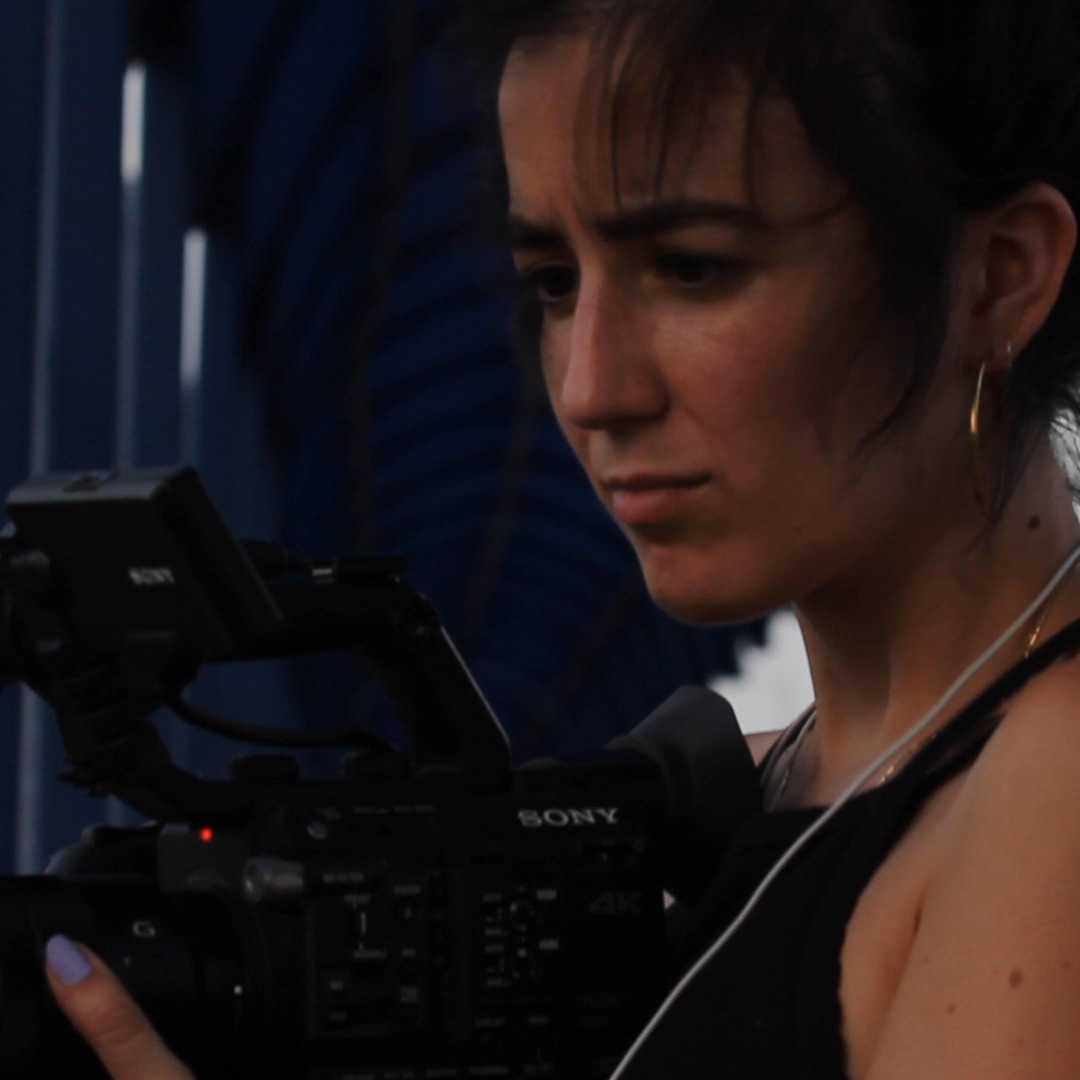 Robie, a Latina woman in a tanktop and her hair pulled back, films on a large Sony camera.