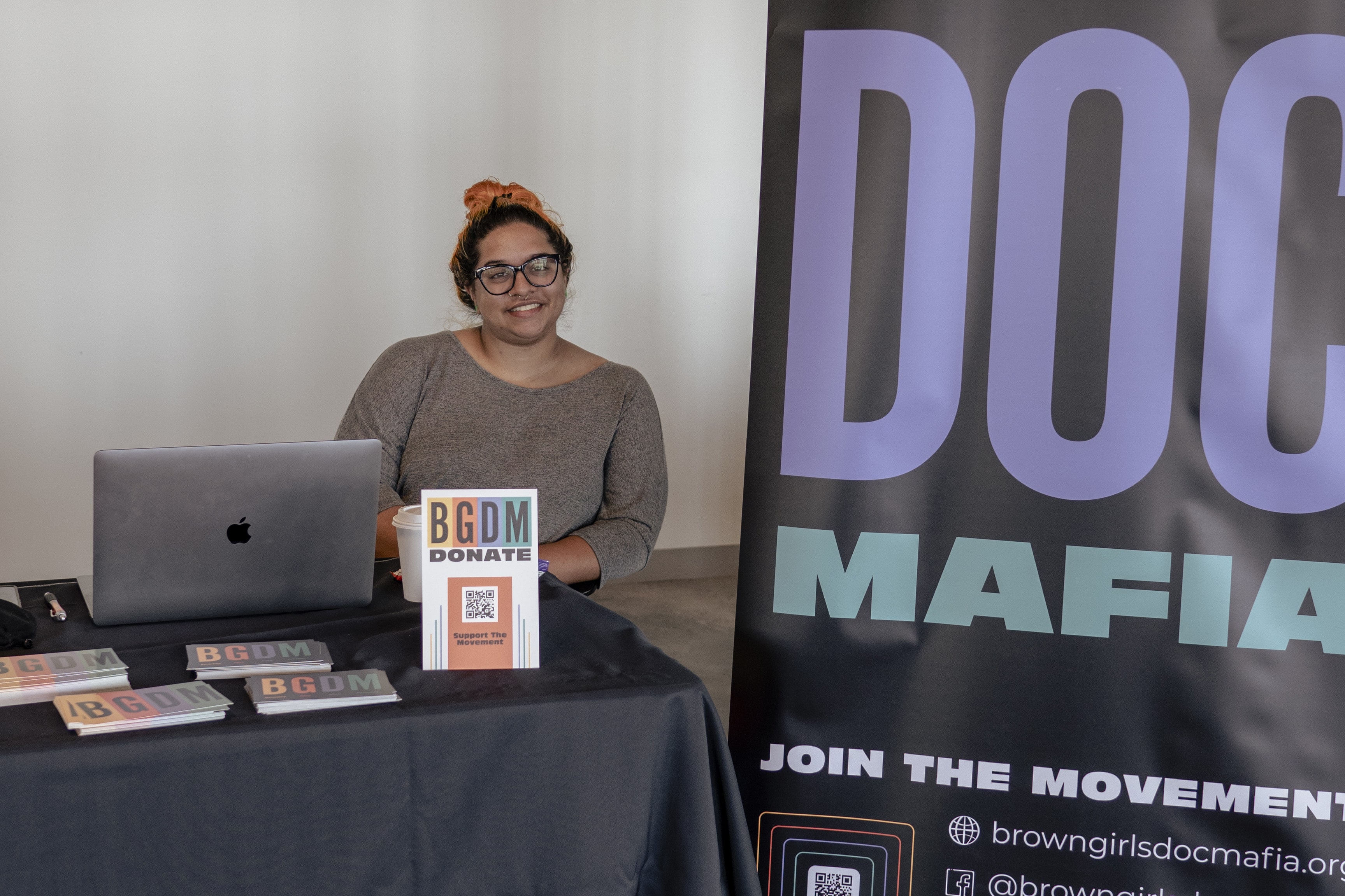 A woman of color with orange hair in a bun sits at a table with a black tablecloth and postcards that say BGDM, next to a large 