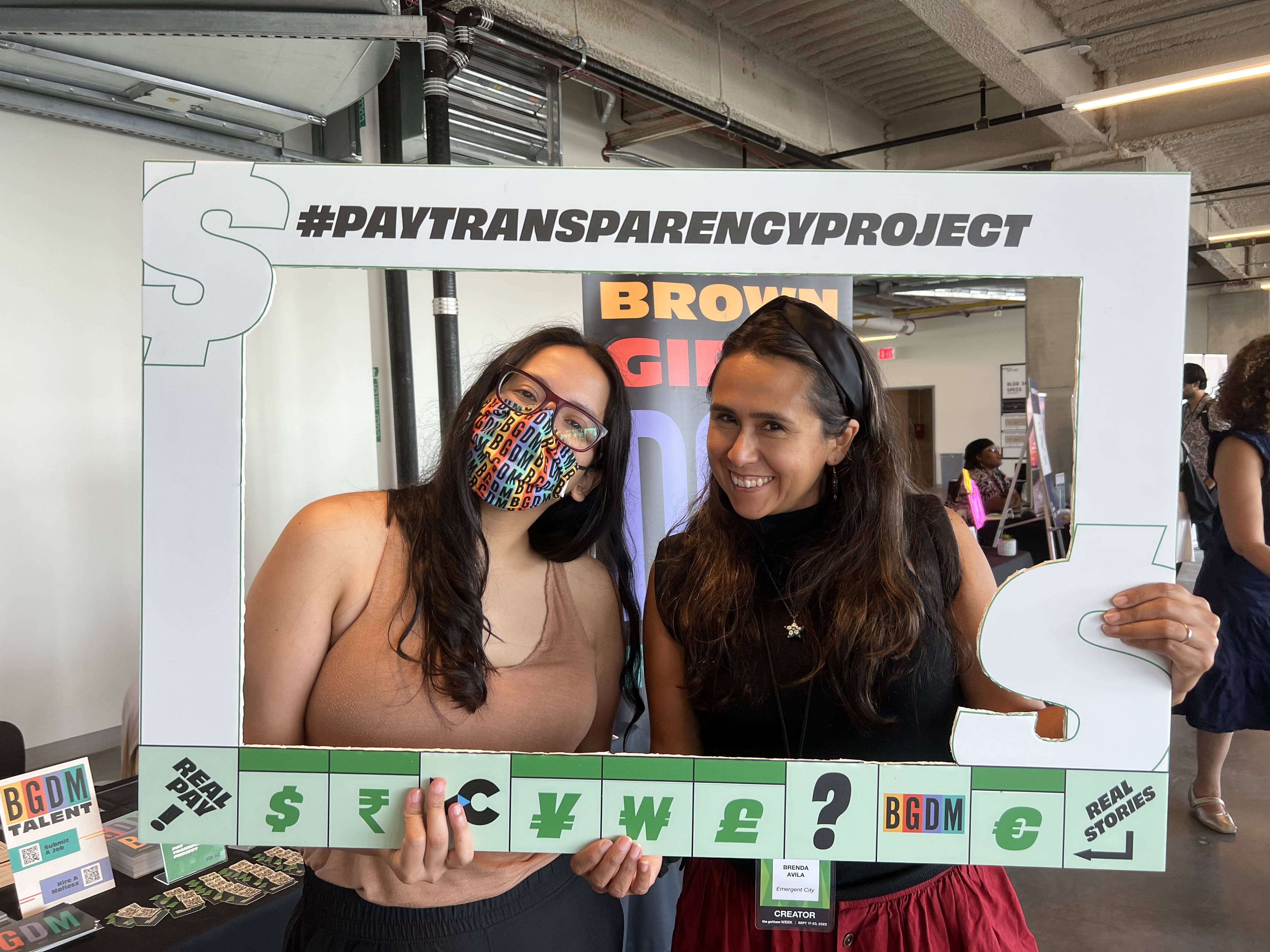 Two women of color with long brown hair pose for a photo, holding up a photo frame around their faces that says Pay Transparency Project, mimicking a Monopoly board. 
