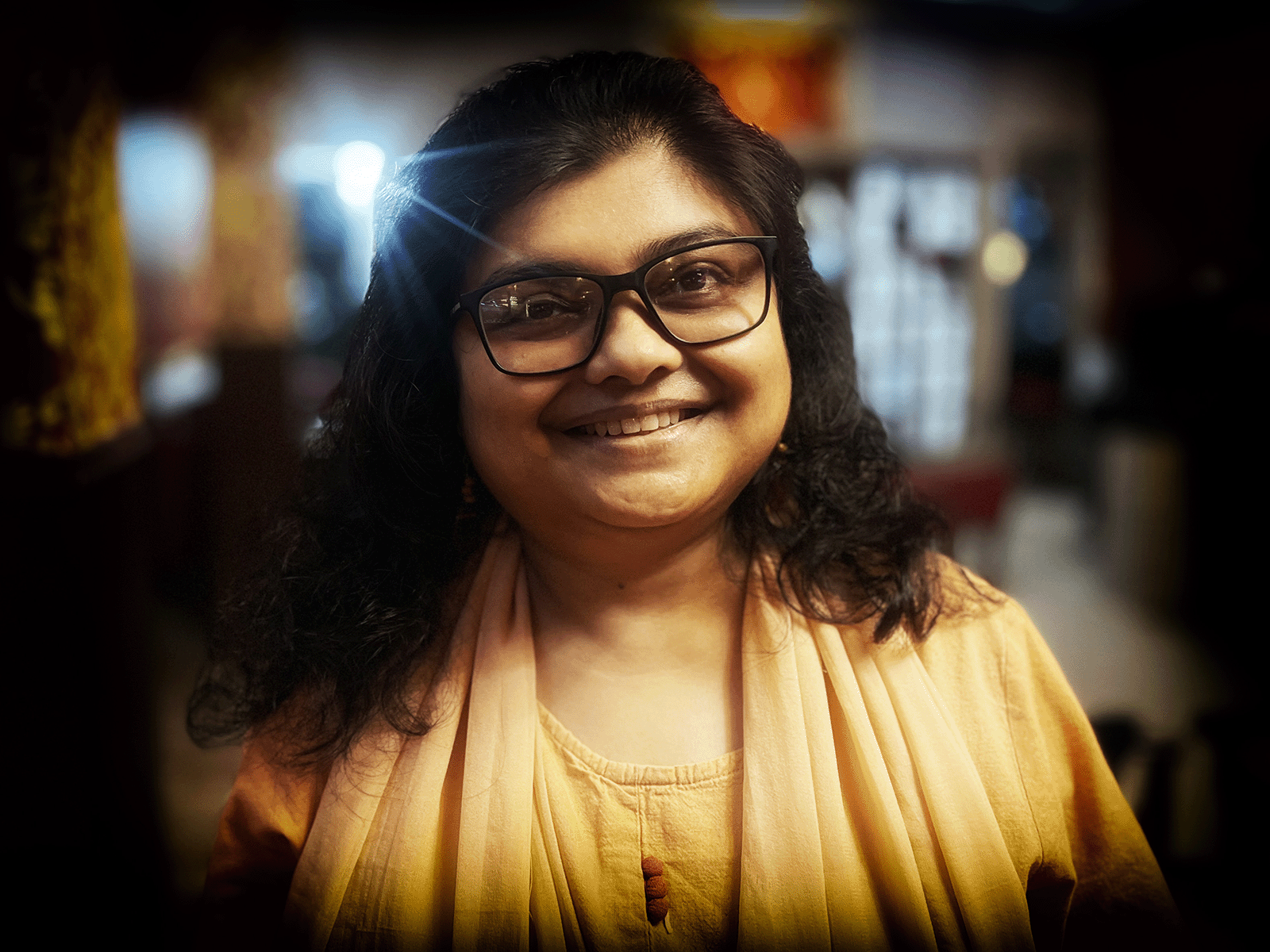 Moupia is an Indian woman with large square glasses, a yellow tunic and wavy brown hair down to her shoulders.