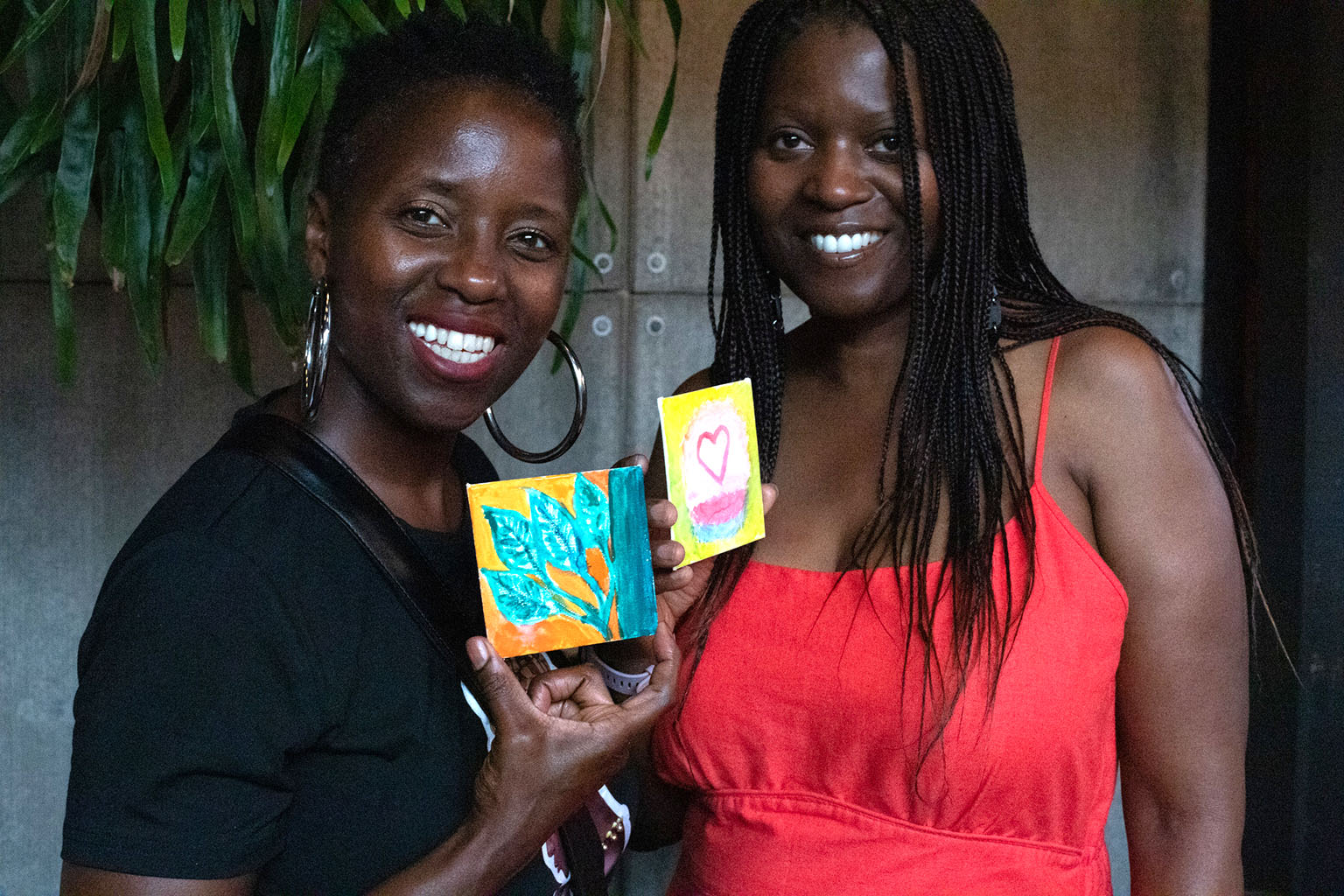 Two Black women smile and pose with their painted square canvases.