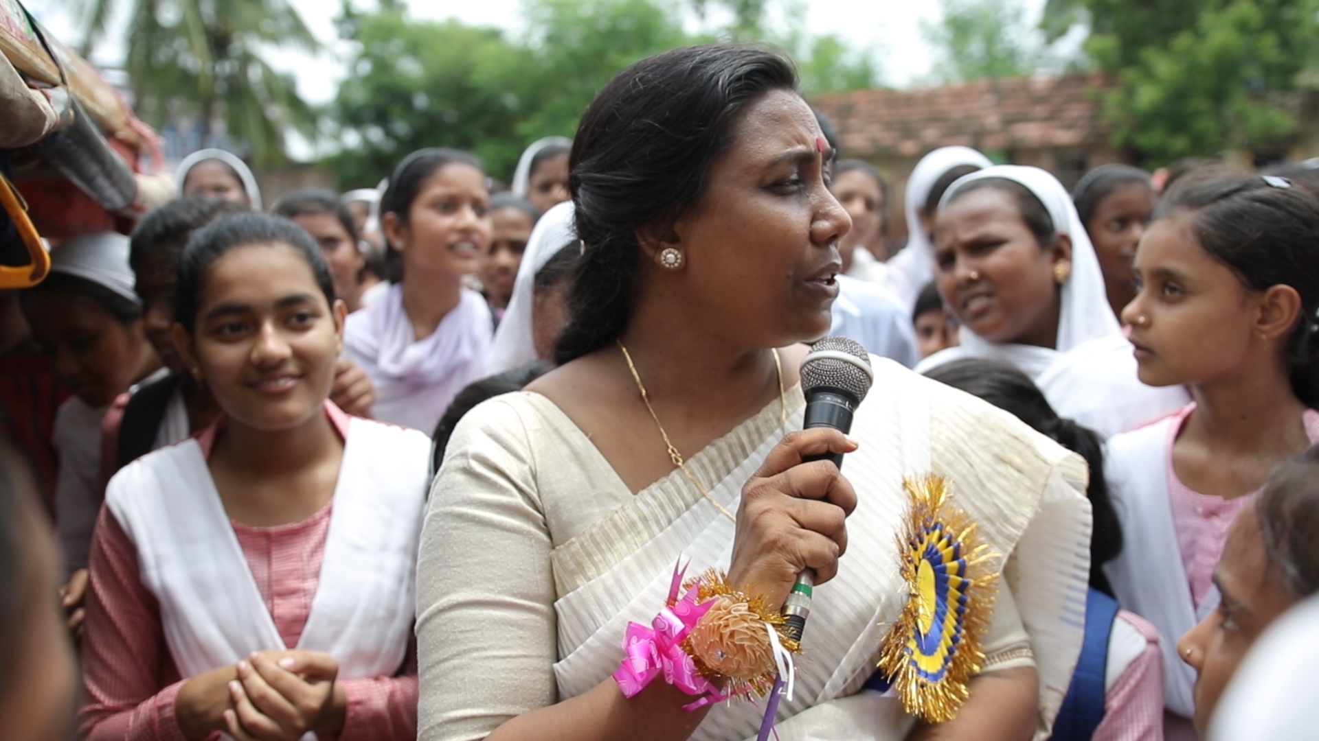 An Indian woman in a beige sari, stands amidst young Indian female students, while holding a microphone.