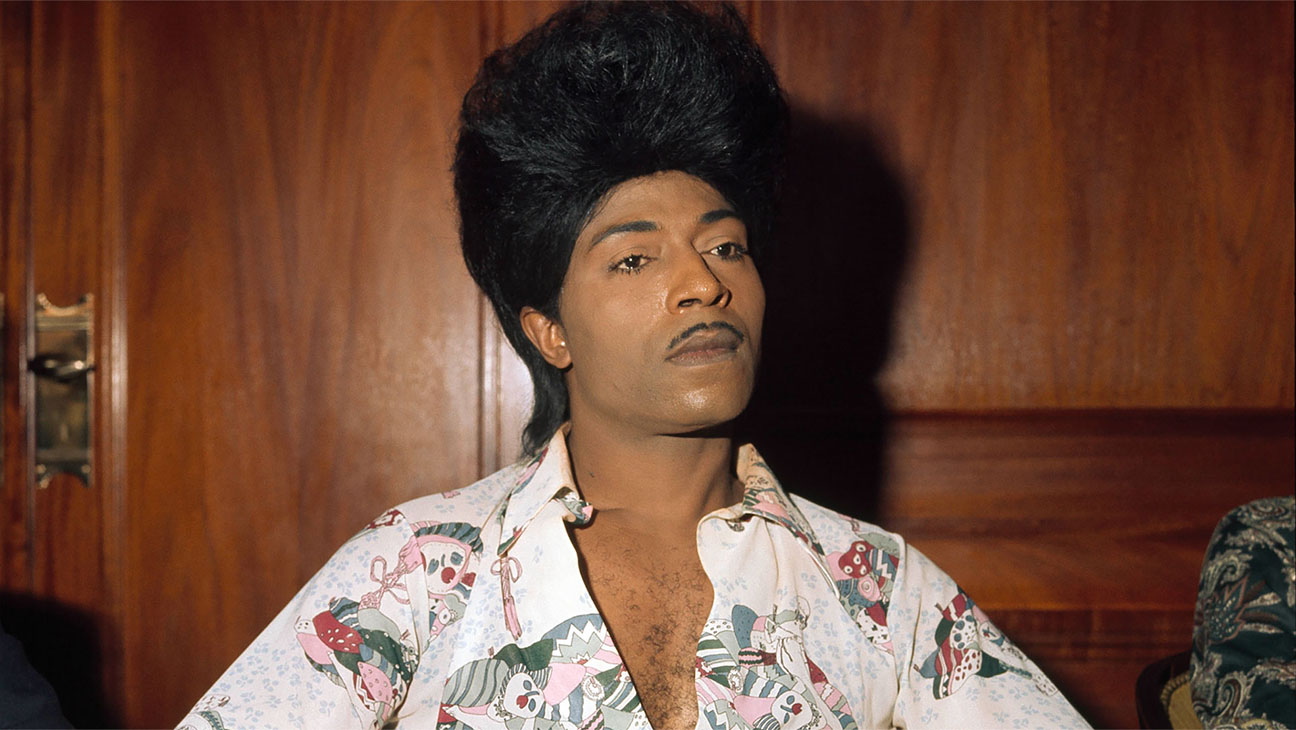 Little Richard, a Black man with a thin moustache, a large coiffed hairstyle a white collared shirt, stands in front of a wood paneled wall.