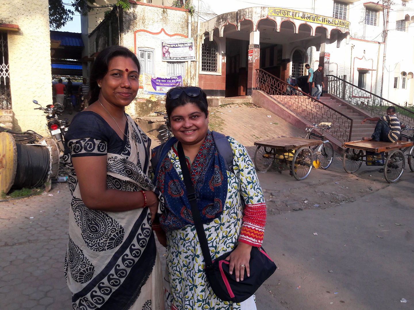 Murshida, a tall Indian woman with long hair in a muted navy blue sari, and Moupia Mukherjee, a short Indian woman with a pixie cut and a patterned kurta, stand in an Indian street.