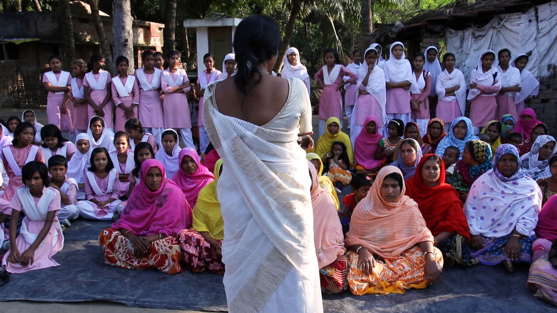 From behind, Murshida, in an ivory sari, addresses the students of her Madrasah, girls dressed in pink and white.