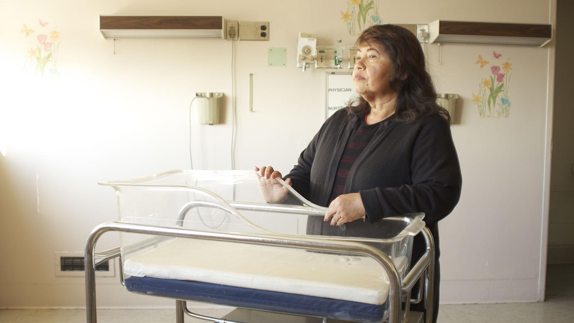 A Latinx woman stands over a hospital baby bassinet looking into the distance