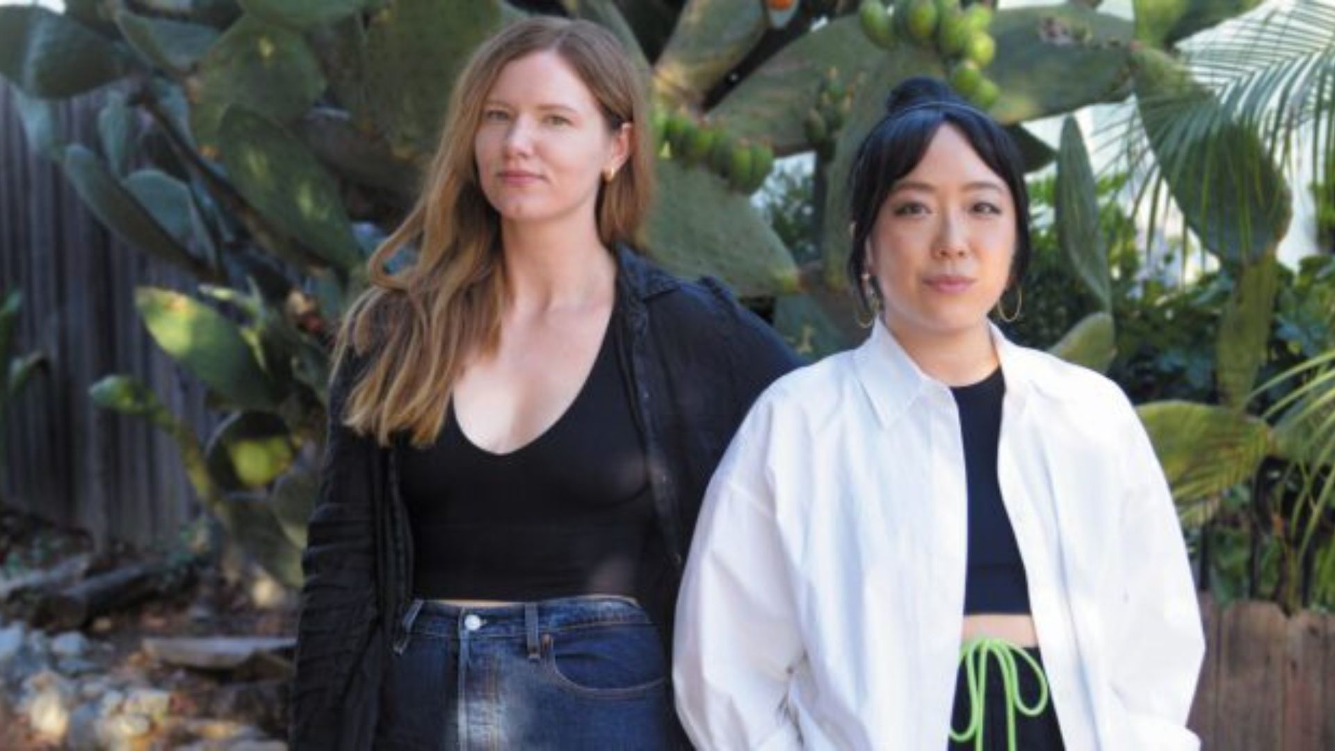 A White woman with long dirty blonde hair and a low-cut, long-sleeve black shirt stands next to an Asian woman with black hair pulled back into a bun, a white open button up and a black shirt underneath. They stand outside in front of a large cactus.