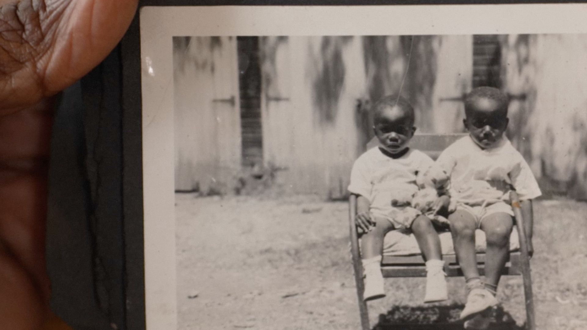 A Black person's hand holds an old black and white photos of two Black babies sitting in a chair.