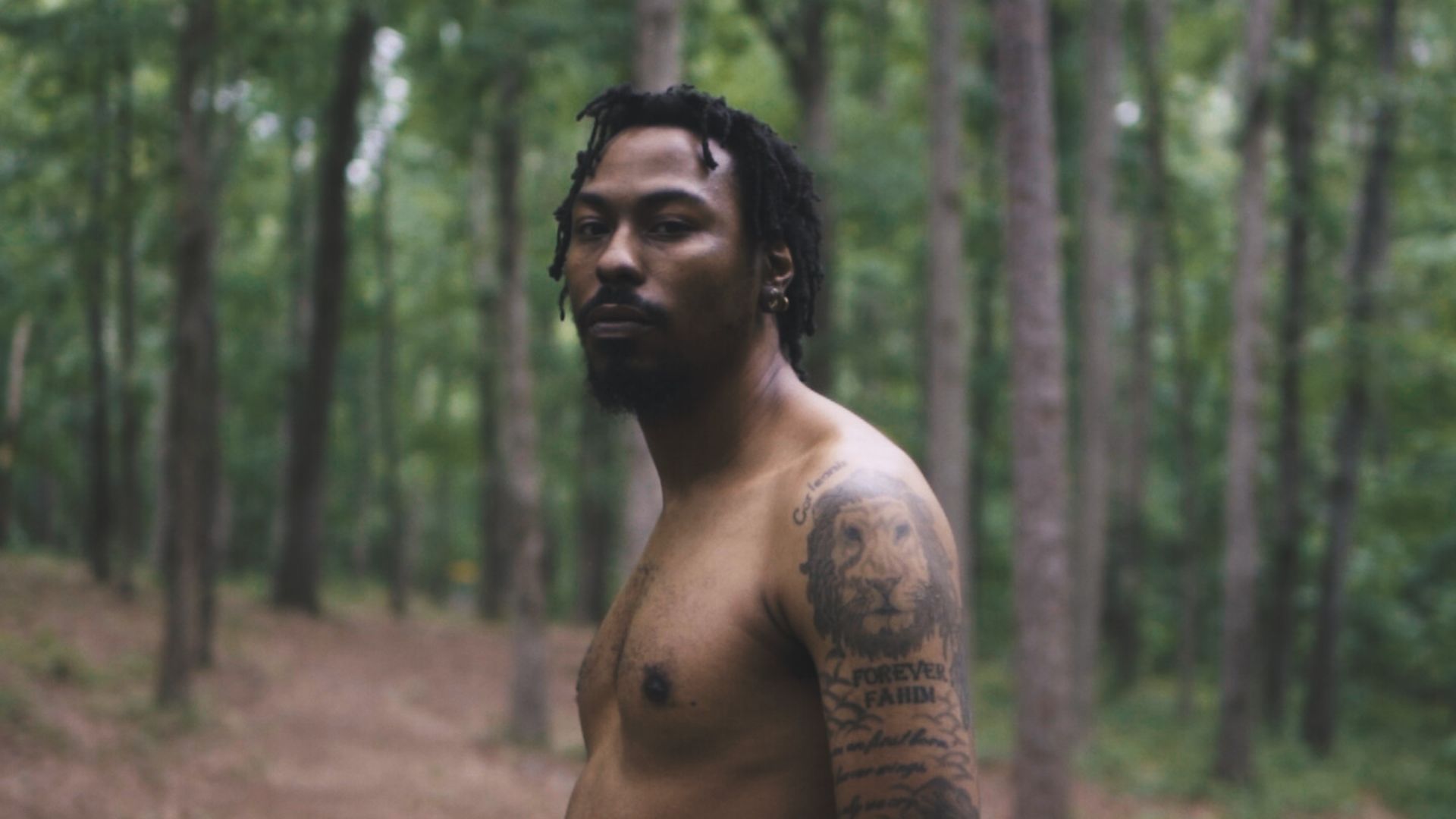 A shirtless tattooed Black man with short dreads stands in the forest, facing the camera.