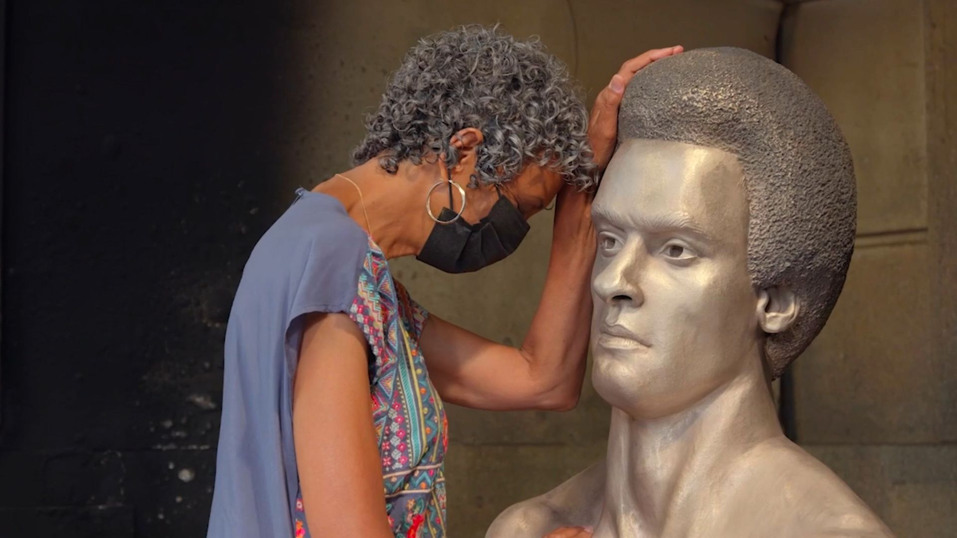 An older Black woman with short curly salt-and-pepper hair, a black face mask and blue shirt rests her hand on and and bows her head in front of a bronze statue of Huey Newton.