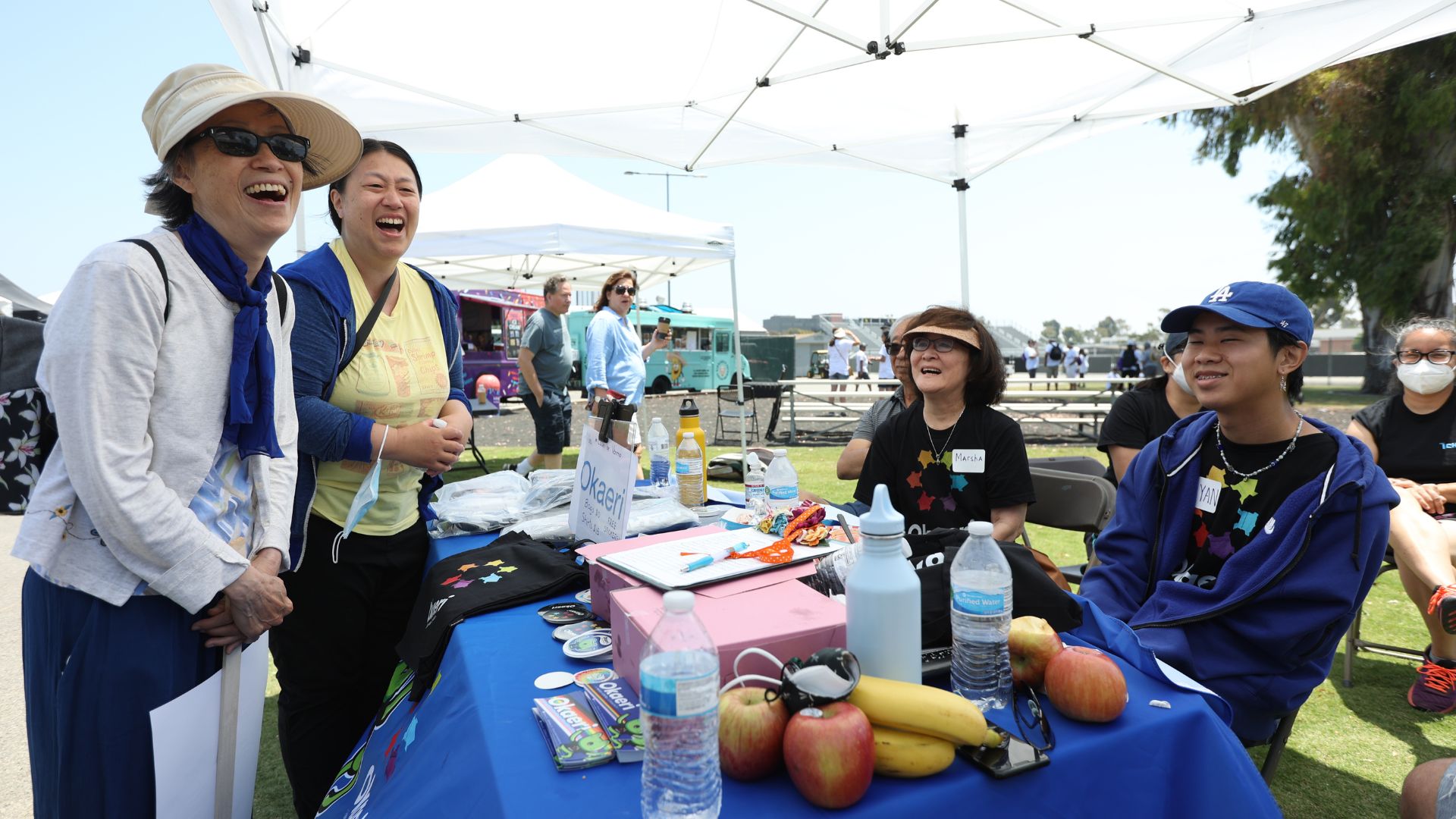 Gathered in a park at a long table with a blue cloth, fruit, shirts, stickers reading "Okaeri." Three older Asian women and a young Asian mad smile and laugh around the table. 