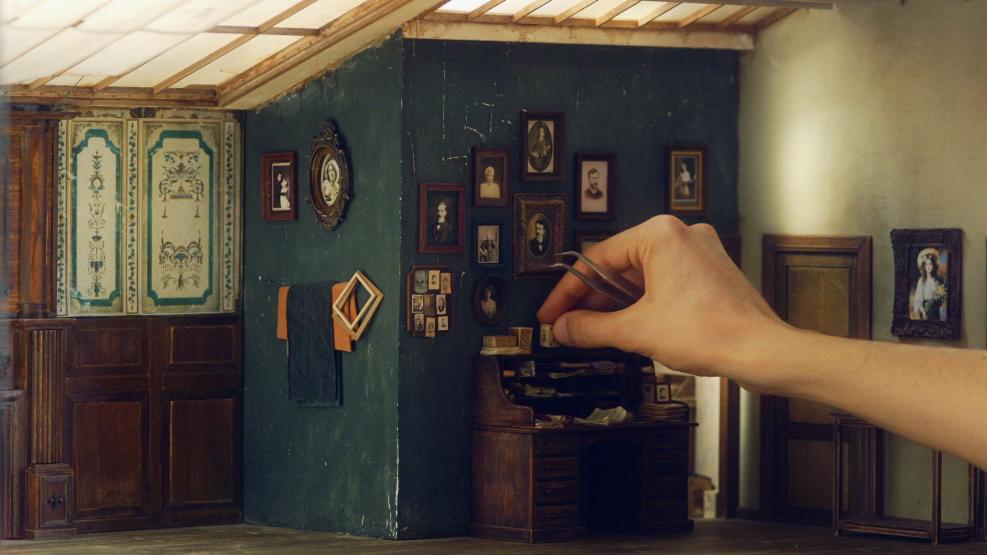 A hand, holding tweezers, places small items onto a desk in a miniature of an old house.