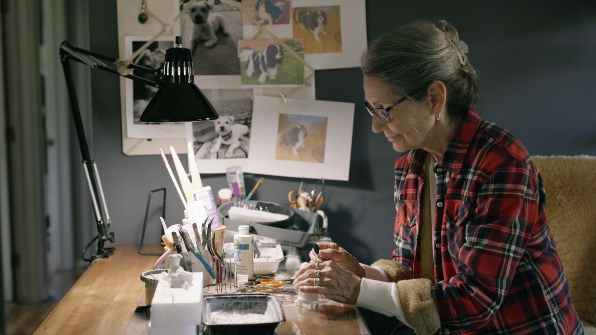 An elderly white woman with grey clipped-back hair, a red flannel shirt and glasses leans over a messy craft table topped with brushes, glue, and paint as she works on creating a miniature white dog. On the wall, printed out papers with photos of dogs. 